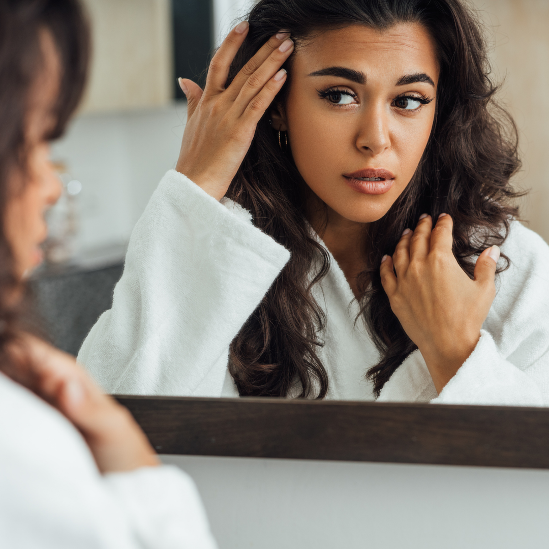 Woman in a white robe lifting her bangs and closely examining her skin and hair in a mirror during pregnancy skincare routine