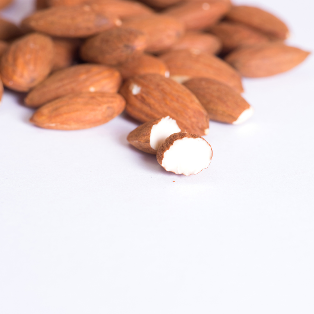 Whole almonds clustered together on a white background with a sliced almond in front revealing the white inner flesh, representing the natural source of mandelic acid